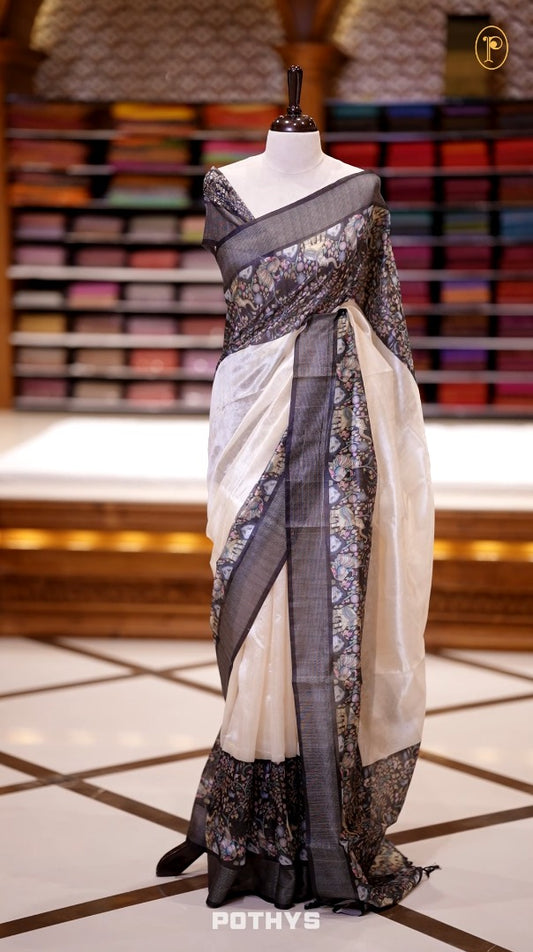 Beige colour semi tussar silk saree featuring with a soft kalamkari & floral prints, accented with a rich black and silver border and elegant printed pallu, paired with a black colour printed with border blouse.