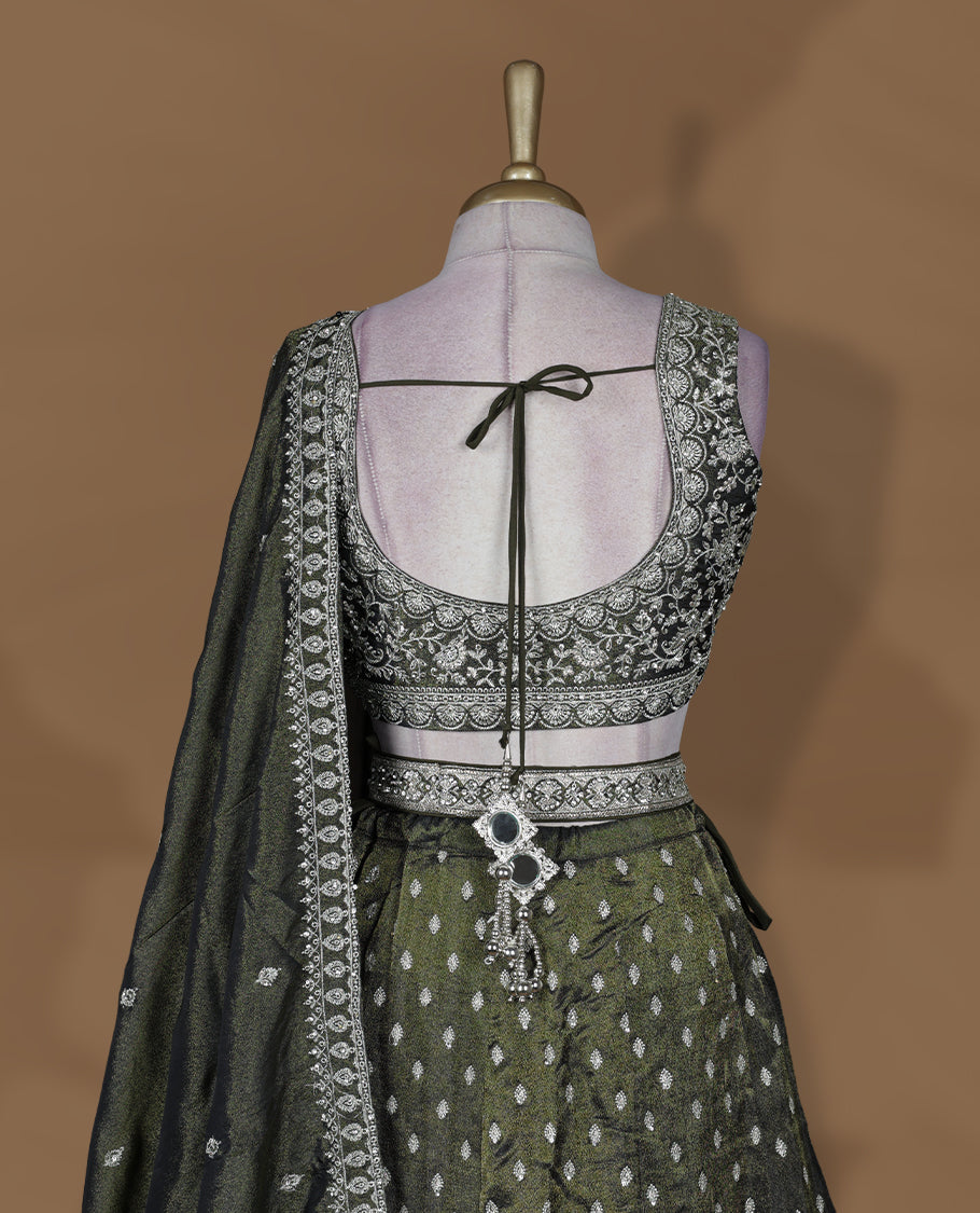 Olive green and black ombre effect lehenga set with stitched sleeveless V‑neck heavily embroidered blouse, matching stitched flared lehenga featuring intricate zari and sequin borders, and coordinated dupatta with embellished edge.