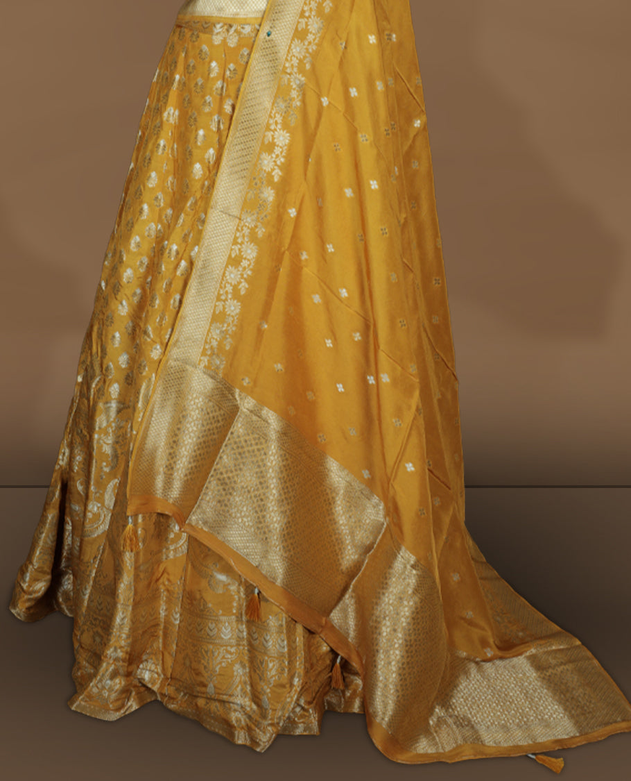 Yellow colour Banarasi lehenga set with a stitched flared lehenga featuring all‑over zari butti motifs and rich woven border, paired with an unstitched floral zari blouse piece and matching dupatta with zari border and tasselled edges.