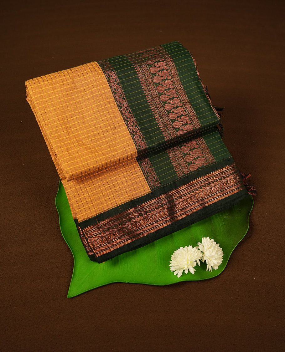 Mustard yellow coloured mixed Kalyani cotton saree featuring copper zari checks on the body, complemented by a dark green coloured border and pallu adorned with copper zari patterns, Paired with a matching blouse.