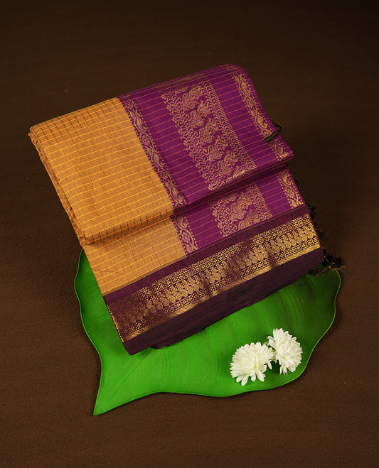 Mustard yellow coloured mixed Kalyani cotton saree featuring golden zari checks on the body, complemented by a grape purple coloured border and pallu adorned with golden zari patterns, Paired with a matching blouse.