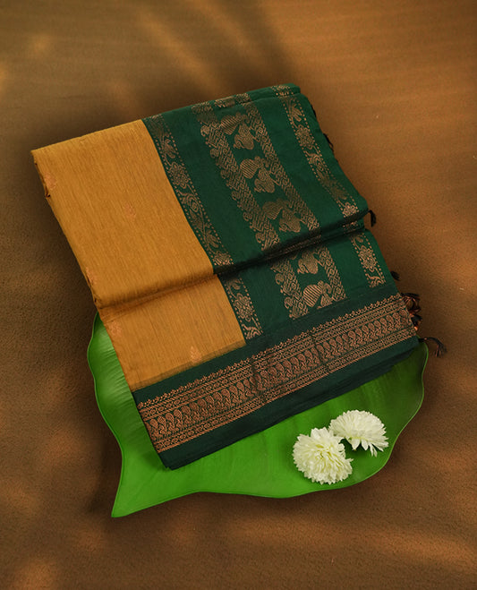 Mustard yellow colour Kalyani Cotton saree with rich green border and pallu, featuring copper zari peacock motifs and intricate weaving & Includes a matching blouse piece.
