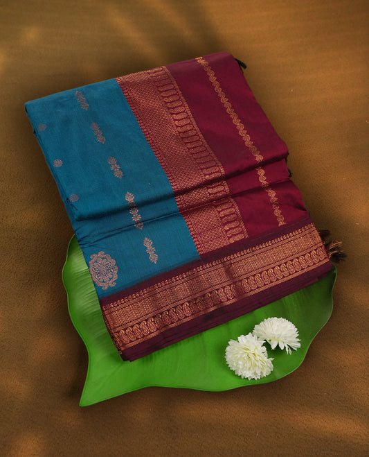 Blue colour Kalyani Cotton saree in deep maroon with golden floral motifs, paired with a contrasting maroon border featuring intricate zari work & Comes with a matching blouse piece.