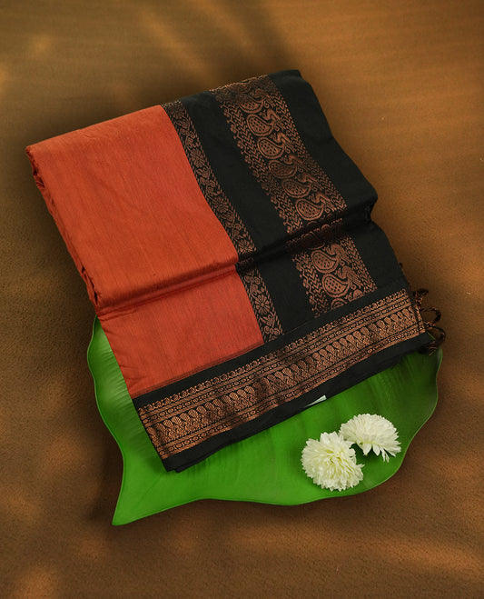 Rust orange colour Kalayani cotton saree highlighted with a black border adorned in intricate copper zari motifs, complemented by a matching pallu and blouse piece.