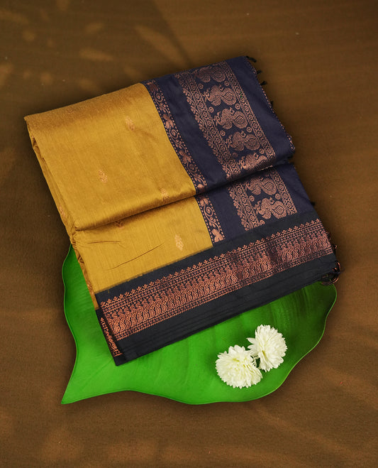 Mustard yellow colour Kalyani Cotton saree with subtle zari woven motifs all over, paired with a contrasting navy blue rich copper zari border & peacock motifs on pallu & Comes with a matching blouse piece.