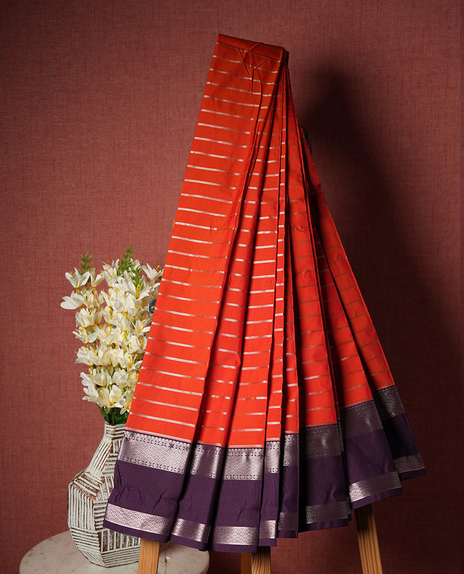 Orange red colour Semi Silk Saree with gold zari striped patterns on the body, paired with a contrasting Dark violet colour traditional pallu and border, and a matching blouse piece.