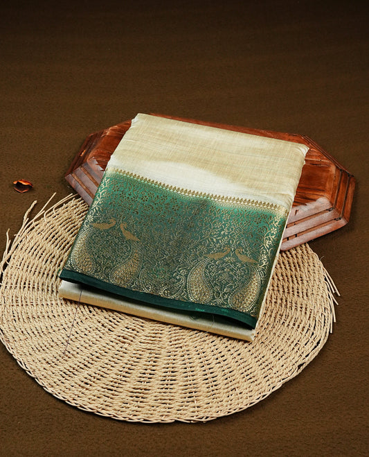 Light gold tussar saree featuring Elegant plain body, complemented by contrast Bottle green colour with Gold zari Floral kodi with Peacock Pattern border, comes with matching blouse piece.