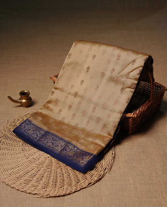 Cream colour Banaras tussar silk saree featuring elegant gold zari buttas across the body, complemented by a contrast Navy Blue border with rich zari detailing. Comes paired with a matching blouse piece.