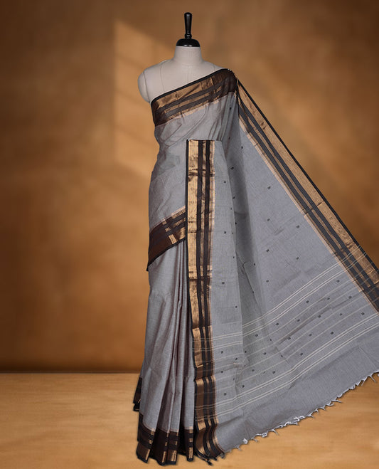 Grey colour chettinad cotton saree featuring small black buttas on body highlighting contrast dark chocolate colour with gold zari traditional border, pallu and paired with matching blouse piece.