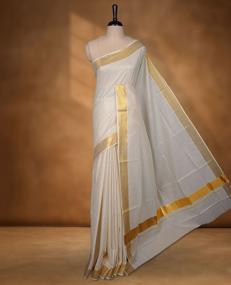 Off-white colour cotton Kerala saree featuring a subtle golden checkered body, highlighted with a classic gold kasavu border accented by fine red lines, and finished with a matching cotton blouse.
