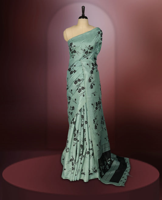 Pastel green colour saree with black floral prints all over the body, complemented by a contrasting black pallu adorned with bold floral designs and striped detailing & Paired with a matching blouse for a complete look.