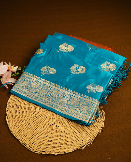 Teal blue colour Banaras Satin Crepe saree featuring gold zari floral motifs on the body, a richly woven traditional border with intricate gold zari detailing, and paired with a matching blouse.