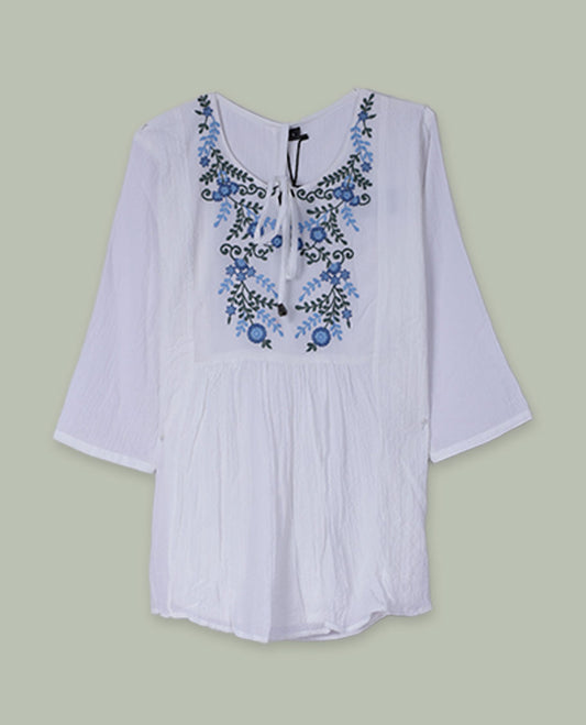 White three-quarter sleeve tunic top with round neckline, front yoke featuring blue and green floral embroidery, and tie-up detail at the neck.