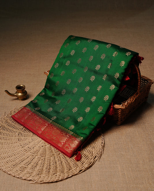 Green colour Banaras tussar silk saree featuring elegant gold zari buttas across the body, complemented by a contrast Red border with rich zari detailing. Comes paired with a matching blouse piece.