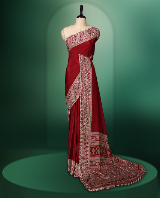 Deep Red colour semi modal saree featuring an ethnic patterned border and pallu in contrasting beige and maroon hues, highlighted with traditional floral motifs, and paired with a matching blouse