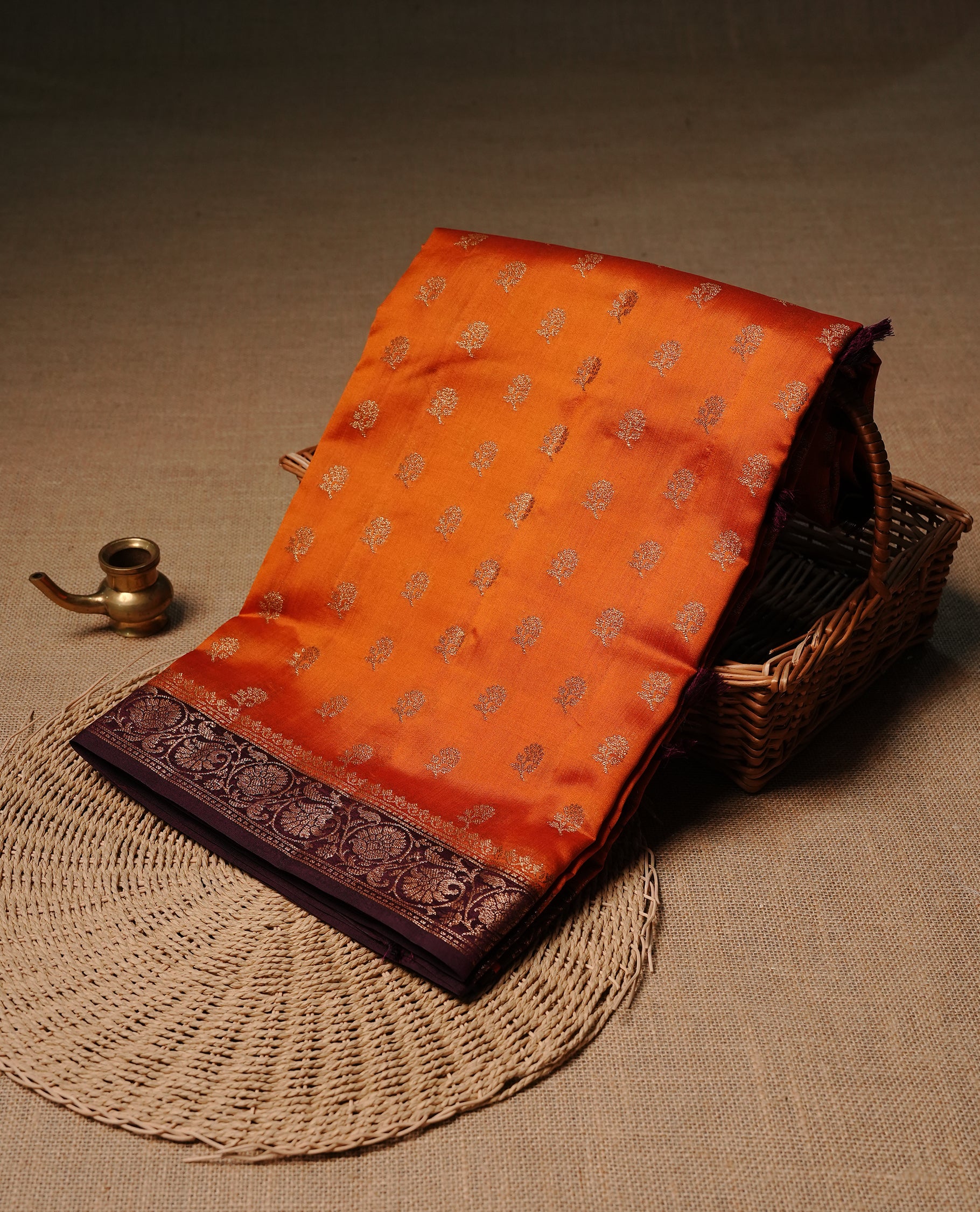 Orange colour Banaras tussar silk saree featuring elegant gold zari buttas across the body, complemented by a contrast chocolate brown border with rich zari detailing. Comes paired with a matching blouse piece.