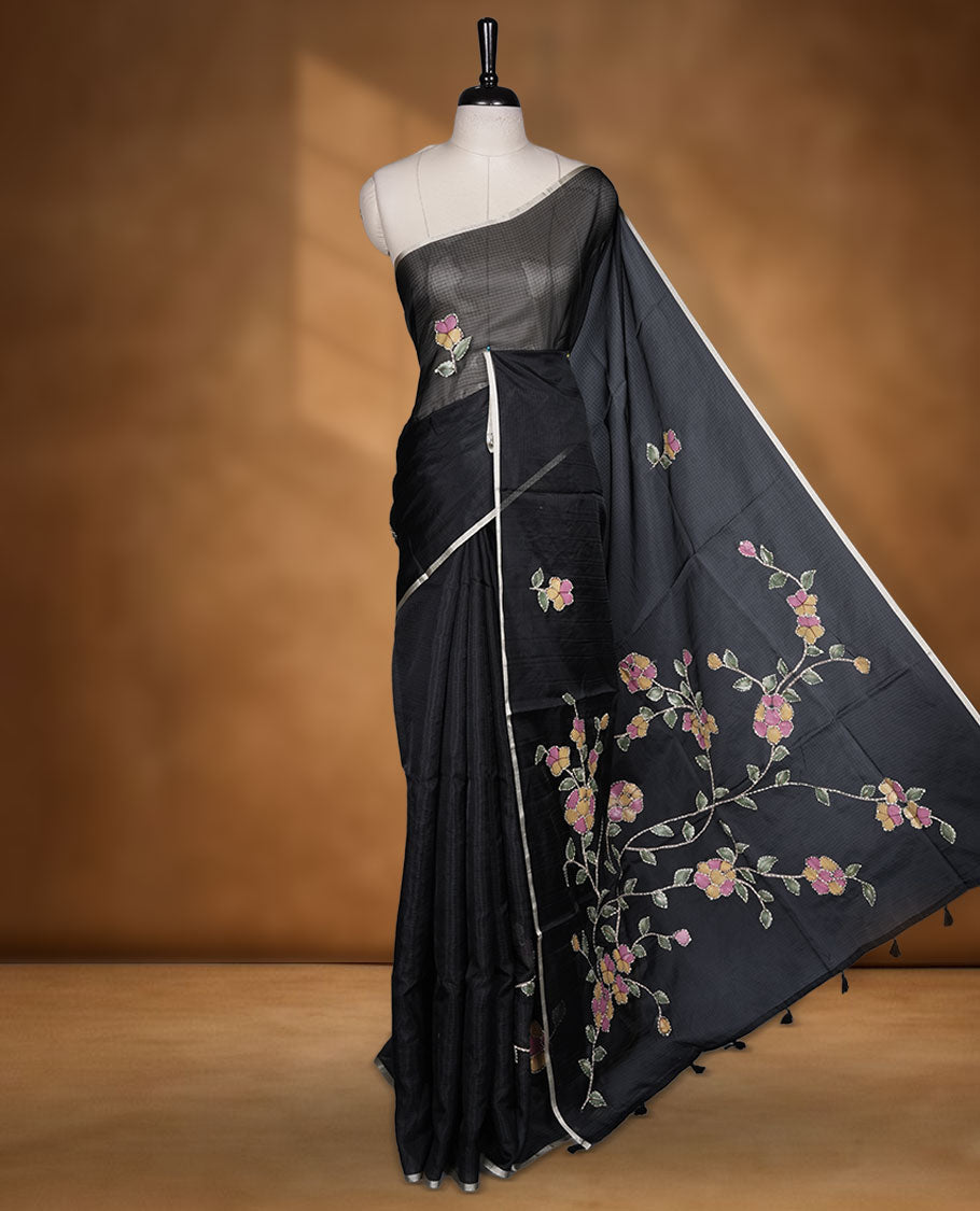 Black colour semi tusser silk saree showcasing delicate embroidered floral motifs throughout the body, finished with soft contrast detailing on the border and complemented by a matching embroidered blouse.