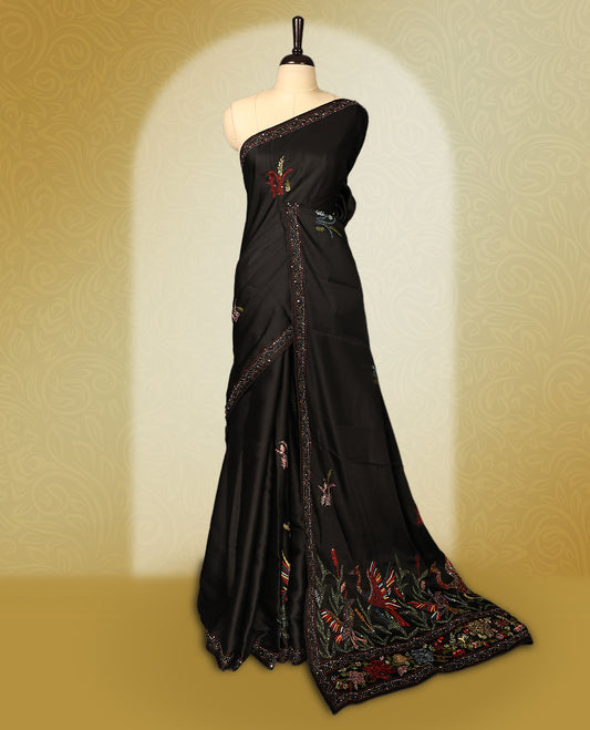 Black colour pure tussar saree adorned with delicate multicolour floral embroidery along the border and scattered motifs across the body, paired with a matching blouse.