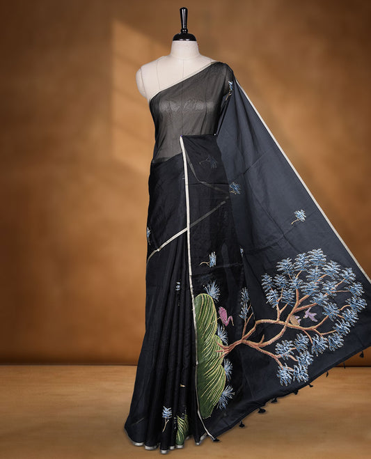 Black colour semi tusser silk saree featuring refined floral embroidery across the body, enhanced with gentle contrast border detailing and accompanied by a perfectly matching embroidered blouse.