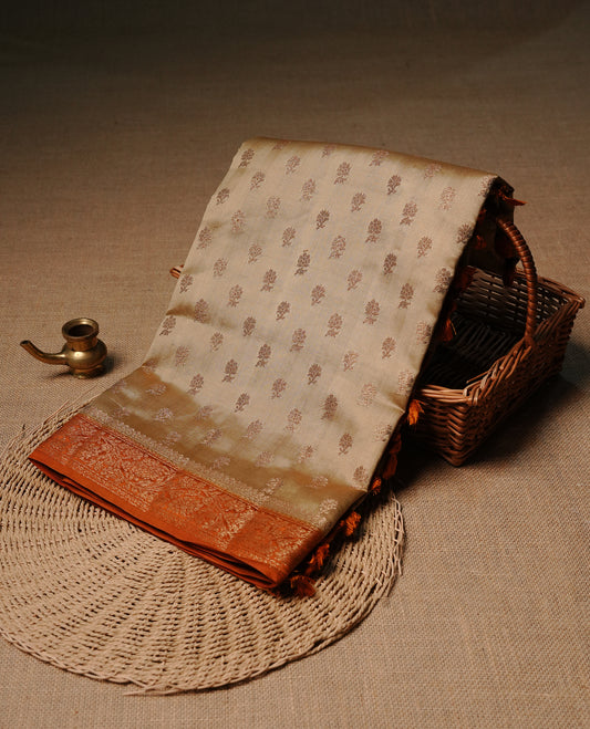 Cream colour pure tussar Banaras silk saree featuring intricate gold zari motifs across the body, enhanced with a contrast orange floral kodi zari border for a rich traditional look. Comes with a matching blouse piece.