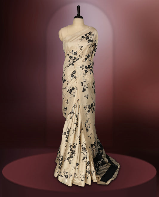 Beige colour saree adorned with black floral prints across the body, complemented by a striking black pallu with floral and stripe detailing. Finished with a subtle border and paired with a matching blouse.