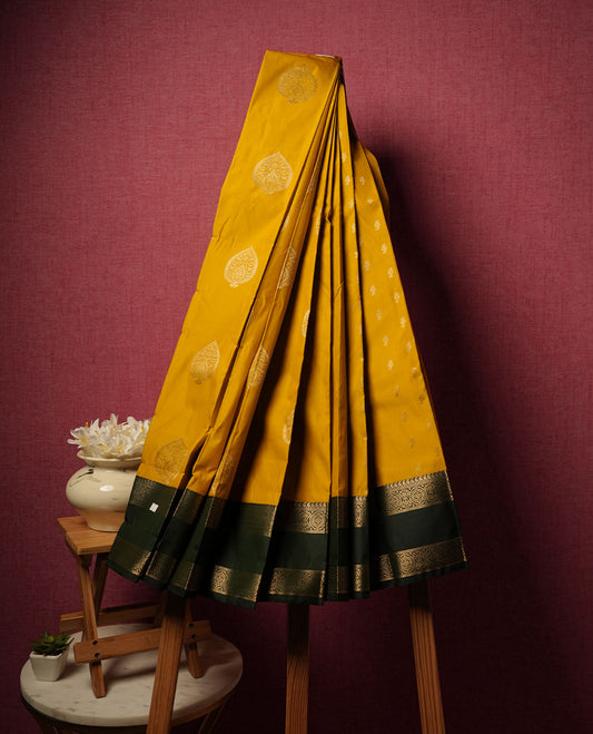 Mustrad yellow colour semi silk saree highlighting gold zari motifs across body, featuring contrast Dark green colour border with gold zari pattern, comes with matching blouse.