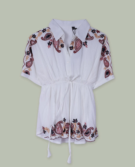 White short tunic top with collared neckline, short sleeves, contrast paisley and floral embroidery on shoulders and hem, and a drawstring waist with tassel ties.