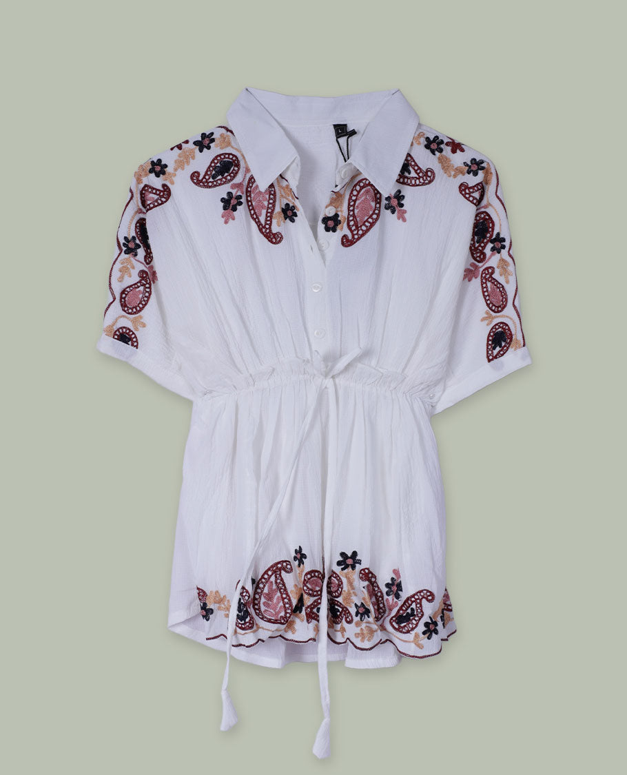 White short tunic top with collared neckline, short sleeves, contrast paisley and floral embroidery on shoulders and hem, and a drawstring waist with tassel ties.