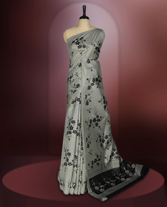 Grey colour saree with black floral prints all over the body, complemented by a contrasting black pallu adorned with bold floral designs and striped detailing & Paired with a matching blouse for a complete look.