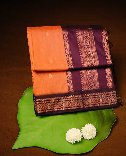 Orange colour kalyani cotton saree featuring gold zari motifs on body, highlighting contrast Purple colour with Antique gold zari border, paired with matching blouse piece.