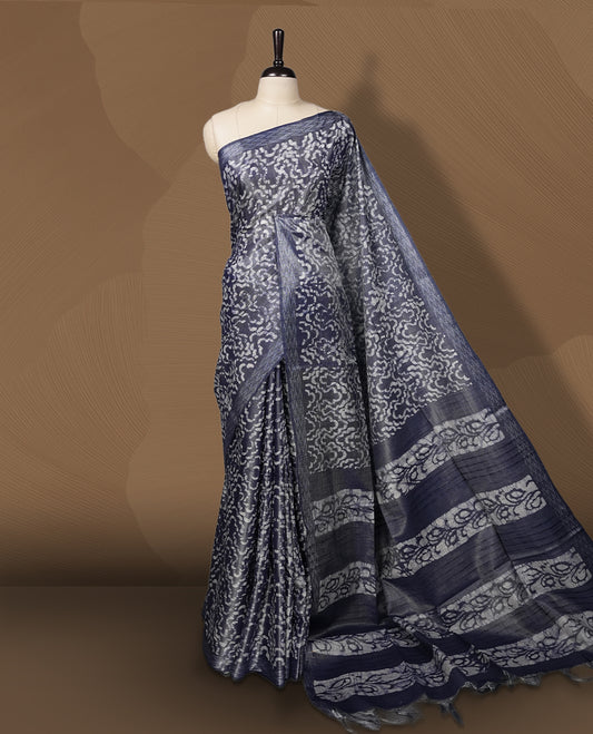 Greyish blue colour semi jute tissue saree featuring white floral prints across the body, adorned with intricate geometric woven borders and a graceful floral work pallu & Comes with a matching blouse piece.