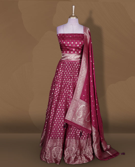 Wine colour Banarasi lehenga set with a stitched flared lehenga featuring all‑over zari butti motifs and rich woven border, paired with an unstitched floral zari blouse piece and matching dupatta with zari border and tasselled edges.