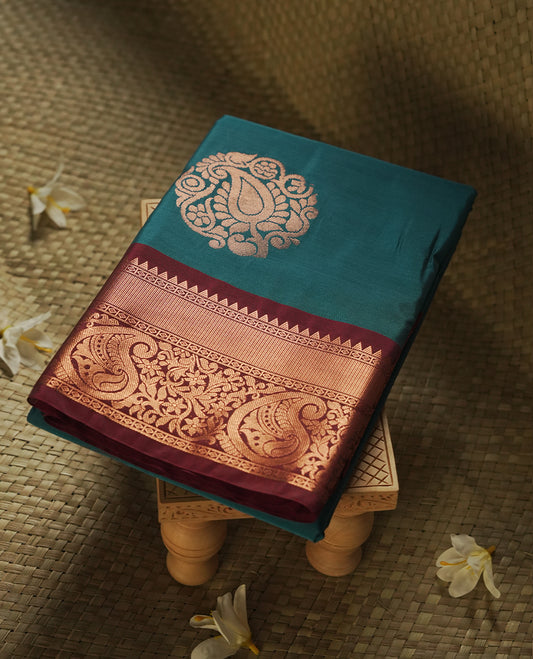 Teal colour Semi Silk Saree with gold zari motifs on the body, paired with a contrast Dark maroon colour pallu with gold zari traditioal border, and a matching blouse piece.