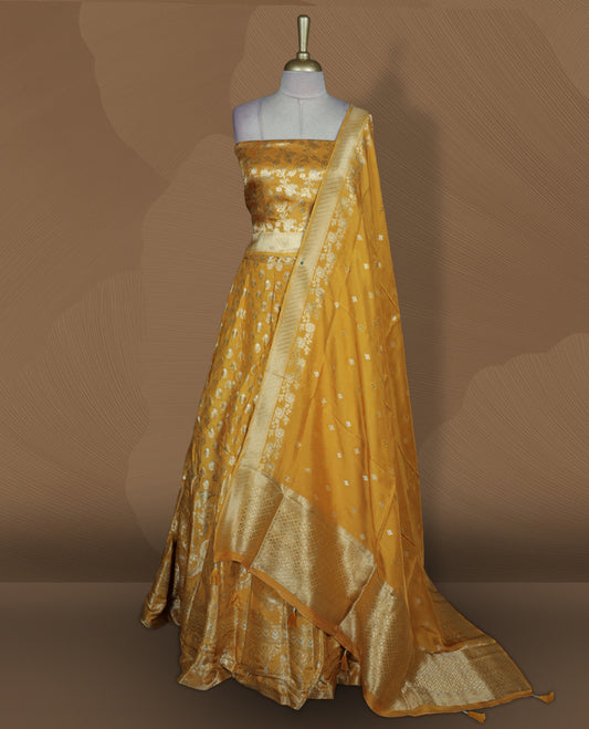 Yellow colour Banarasi lehenga set with a stitched flared lehenga featuring all‑over zari butti motifs and rich woven border, paired with an unstitched floral zari blouse piece and matching dupatta with zari border and tasselled edges.