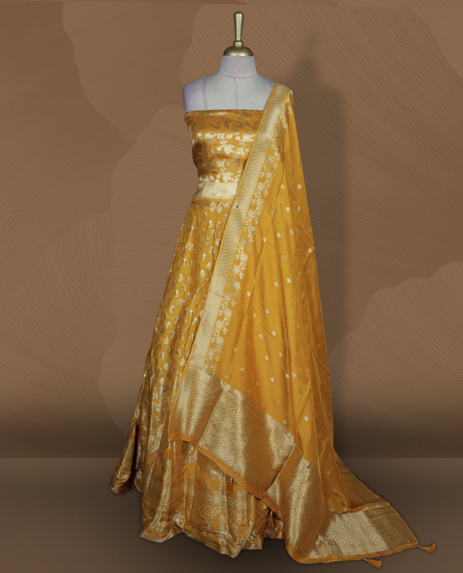 Yellow colour Banarasi lehenga set with a stitched flared lehenga featuring all‑over zari butti motifs and rich woven border, paired with an unstitched floral zari blouse piece and matching dupatta with zari border and tasselled edges.