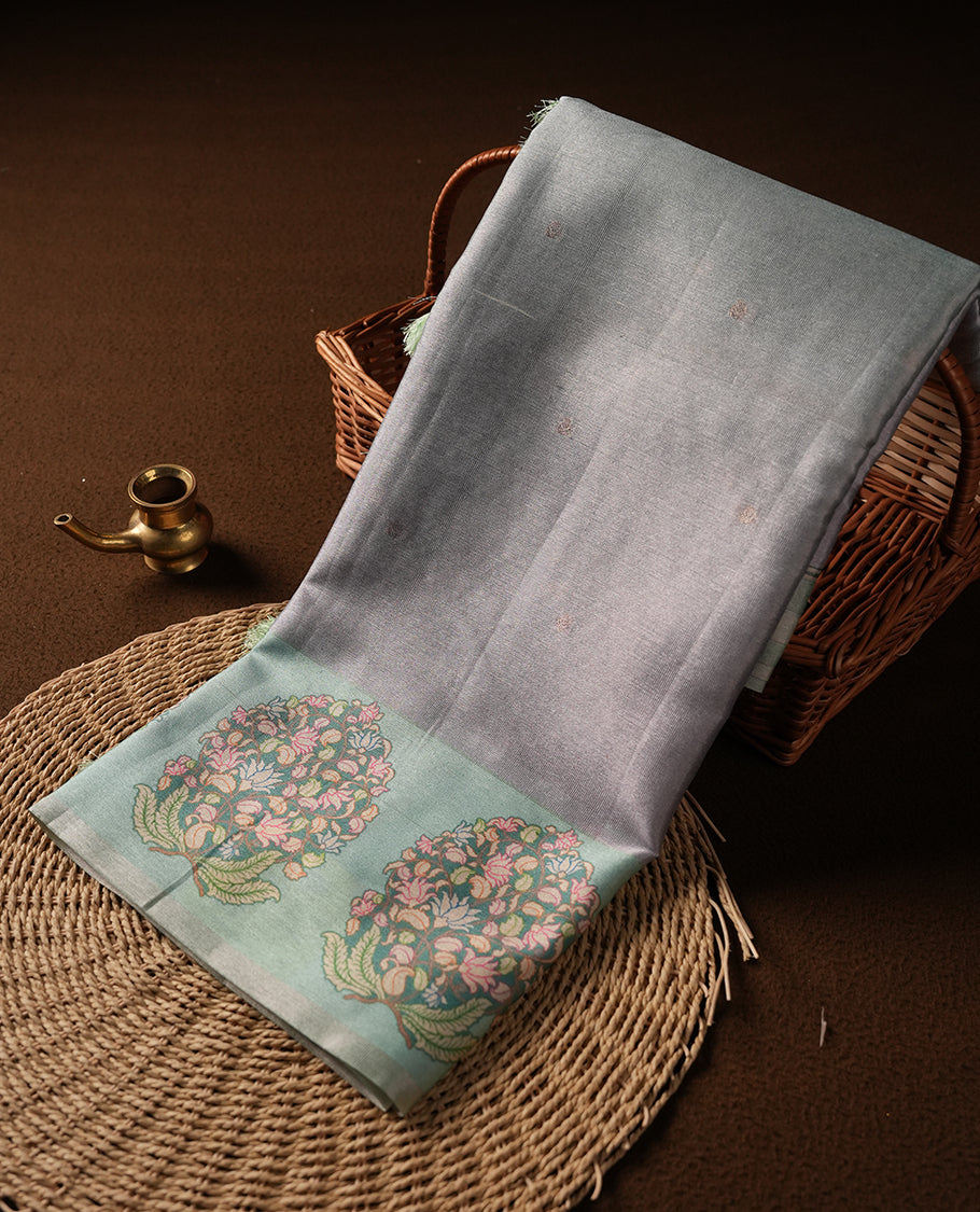 Silver colour semi tussar saree adorned with gold zari motifs across the body and Floral pattern contrast Aqua green colour on border, paired with a matching blouse.