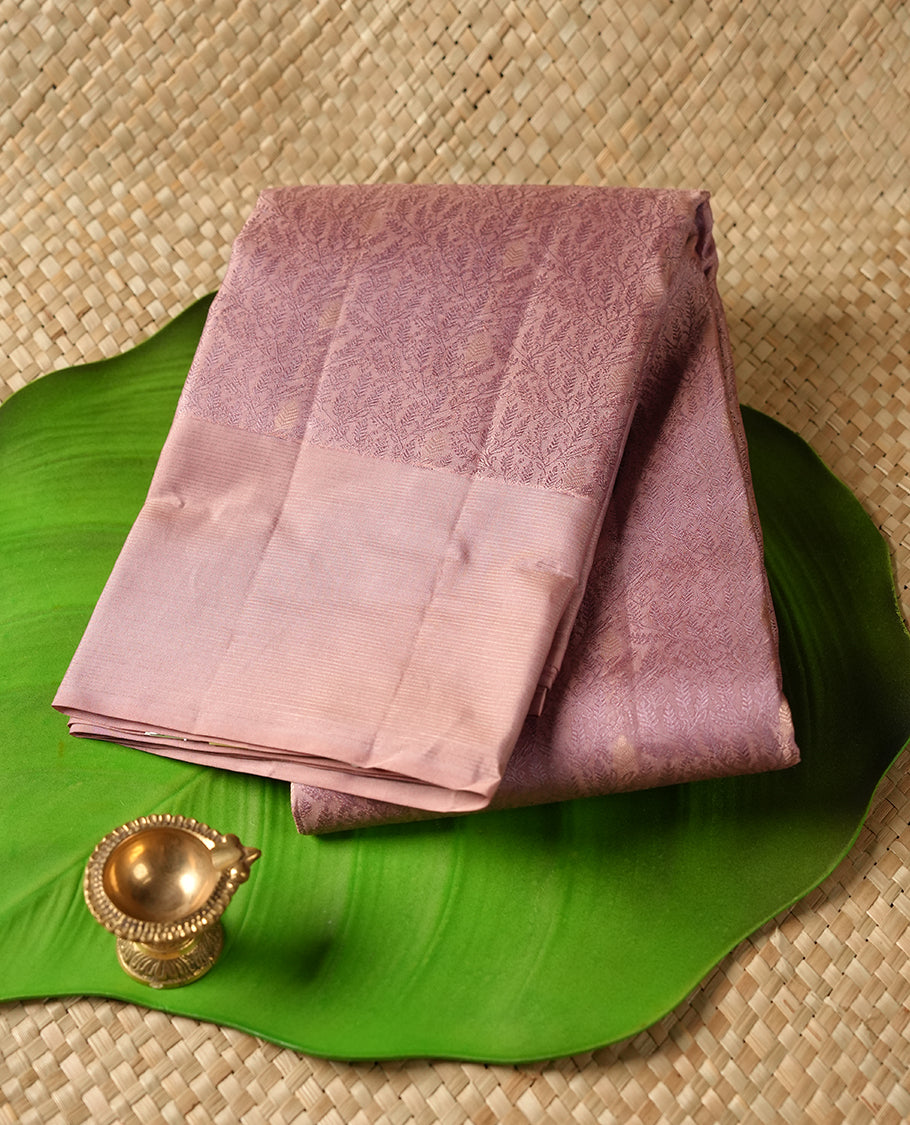 Dusty lavender colour Vasundhara silk saree adorned with all-over self color leaf kodi motifs on the body, complemented by a self colour with gold line pattern border and paired with a matching blouse piece.
