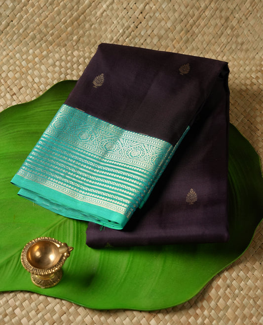 Dark purple colour Vasundhara silk saree adorned with all-over gold motifs on the body, complemented by a contrast green with gold tarditional border and paired with a matching blouse piece.
