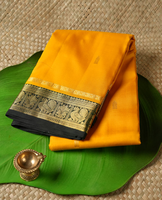 Mustard Yellow colour Vasundhara silk saree adorned with all-over gold motifs on the body, complemented by a contrast dark green colour with gold mayil motifs border and paired with a matching blouse piece.