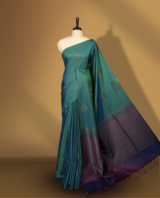 Dual tone blue green semi soft silk saree adorned with woven circular motifs on the body, complemented by a rich contrast plum colour woven pallu with tassel detailing & Comes with a matching blouse piece.