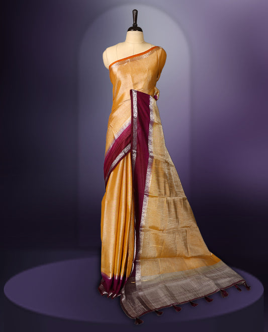 Golden yellow saree with a rich maroon border and pallu, accentuated by delicate silver zari detailing and tassel finish & The fabric has a lustrous sheen and comes with a matching blouse piece.