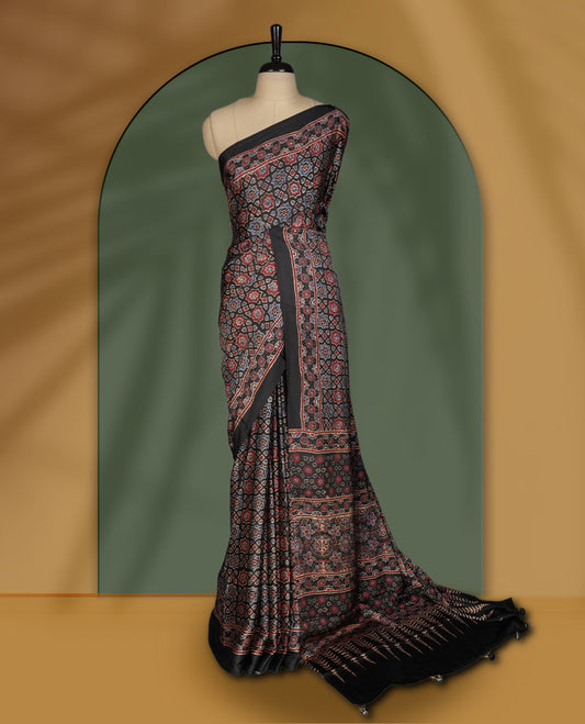 Black colour saree featuring intricate traditional prints in earthy tones, finished with a sleek satin border and tassel details & Comes with a stylish matching blouse.
