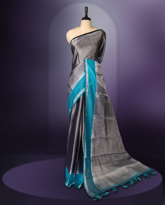 Steel grey saree with a rich sheen, paired with a contrasting turquoise blue border and pallu featuring delicate silver zari work, finished with tassels at the edge. Comes with a matching blouse piece.