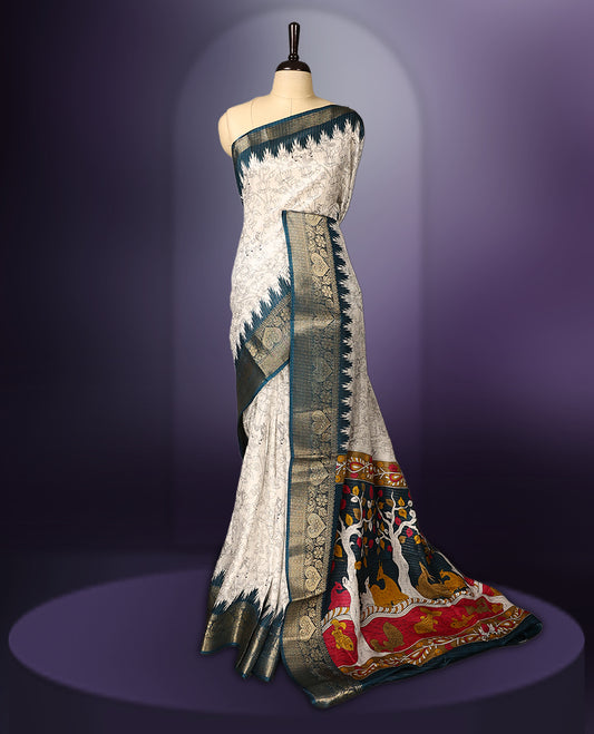 White colour saree adorned with delicate floral designs across the body, enhanced by a rich teal and gold zari border, featuring a vibrant artistic pallu with tree and animal motifs, paired with a matching blouse.