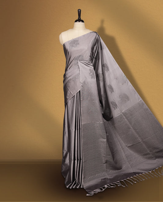 Grey colour semi soft silk saree featuring intricate woven floral motifs on the body, complemented by a rich woven border and pallu with tassel finish & Comes with a matching blouse piece for an elegant look.
