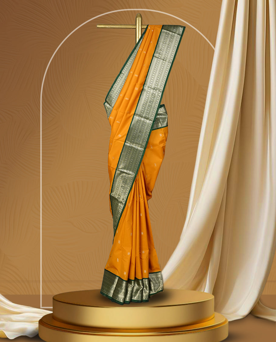 Mustard yellow colour soft silk saree adorned with golden buttas, highlighted by a rich green border and pallu with intricate zari designs & Paired with a matching blouse.