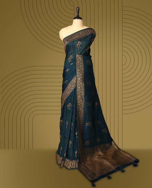 Dark teal green semi Banaras tussar saree adorned with golden floral motifs along the border and scattered designs across the body, finished with a rich golden zari pallu, paired with a matching blouse.