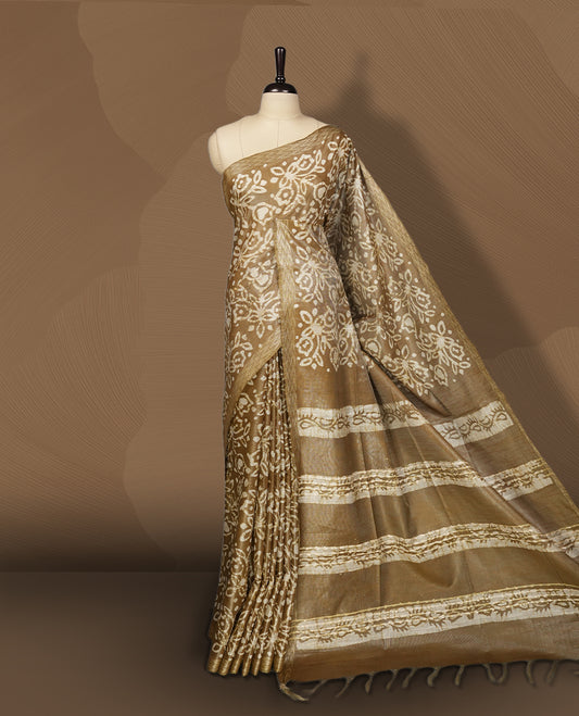 Golden brown semi jute tissue saree featuring elegant floral motifs all over the body, complemented by a shimmering zari striped pallu, woven golden border, and matching blouse piece for a graceful look.