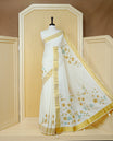 Cream Colour Cotton Kerala Saree Featuring Digital Floral Prints Across The Body, Enhanced With A Gold Zari Border, Traditional Gold Zari Pallu, Finished With Light Brown & White Tassels And A Matching Blouse Piece.