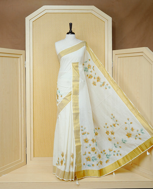 Cream Colour Cotton Kerala Saree Featuring Digital Floral Prints Across The Body, Enhanced With A Gold Zari Border, Traditional Gold Zari Pallu, Finished With Light Brown & White Tassels And A Matching Blouse Piece.